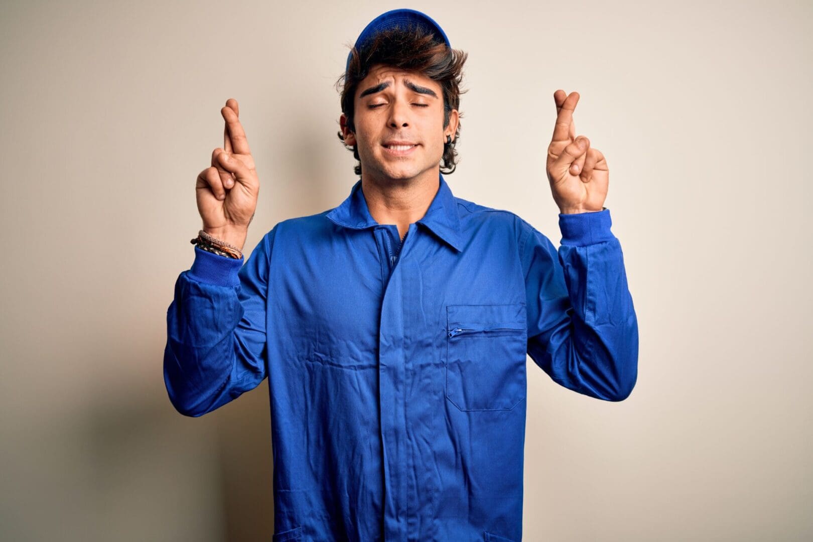 Young mechanic man wearing blue cap and uniform standing over isolated white background gesturing finger crossed smiling with hope and eyes closed. Luck and superstitious concept.