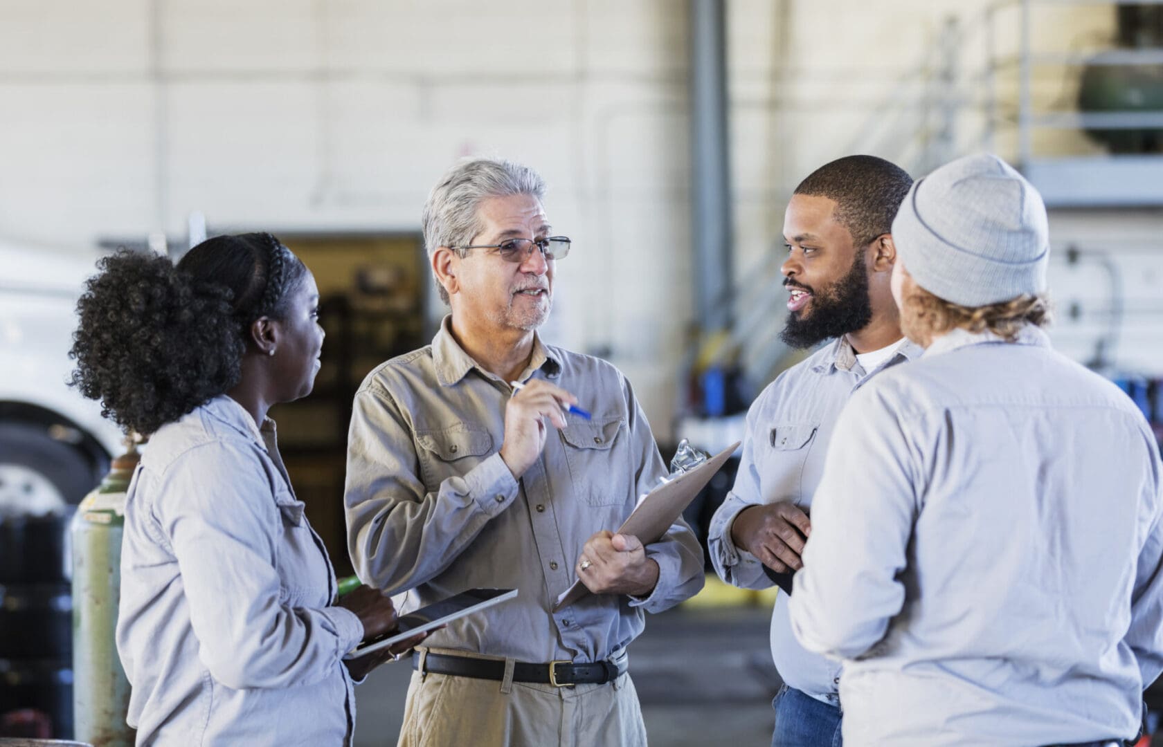 A multi-ethnic group of four workers in a repair shop, standing together, having a meeting. The senior Hispanic man in his 60s, holding the clipboard, is in charge, directing the others. An African-American woman in her 30s is part of the group, listening to the manager, holding a digital tablet.